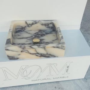 Puede incluir: Un lavabo cuadrado de m&aacute;rmol con un patr&oacute;n veteado crema y gris. El lavabo tiene un desag&uuml;e dorado y se exhibe sobre un pedestal blanco. Las palabras "NATURAL MARBLE" son visibles en el pedestal.