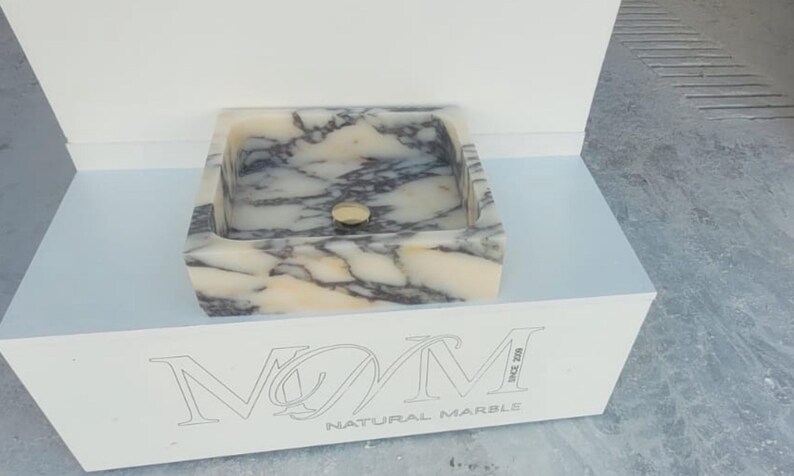 Puede incluir: Un lavabo cuadrado de m&aacute;rmol con un patr&oacute;n veteado crema y gris. El lavabo tiene un desag&uuml;e dorado y se exhibe sobre un pedestal blanco. Las palabras "NATURAL MARBLE" son visibles en el pedestal.