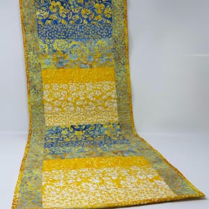 May include: A rectangular, quilted table runner with a patchwork design. The runner features alternating bands of blue and yellow fabric with floral patterns. The edges are finished with a brown border. The runner is approximately 60 inches long.