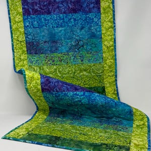 May include: A rectangular fabric table runner featuring horizontal strips of floral and leaf-patterned fabric in shades of purple, blue, teal, and green. The runner is bordered by a bright lime green fabric with a repeating leaf pattern.