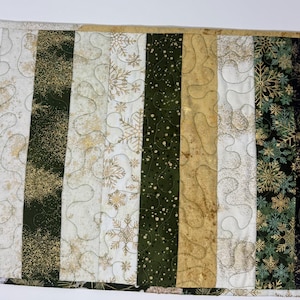 May include: A rectangular quilted table runner featuring vertical fabric strips in shades of forest green, cream, gold, and black. The design includes patterns of pine branches, snowflakes, and gold speckles, finished with a meandering stitch.