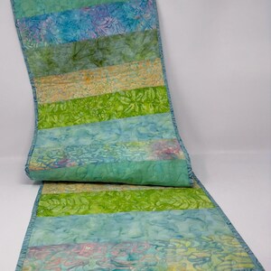 Puede incluir: Camino de mesa acolchado con rayas de tela batik verde, azul y amarilla. Los bordes son de un color verde azulado más oscuro. El camino de mesa mide aproximadamente 30 cm de ancho y 102 cm de largo.