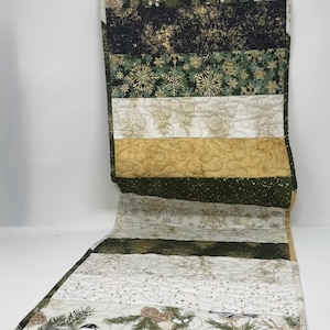 May include: A rectangular quilted table runner featuring horizontal bands of fabric with winter-themed patterns. The design includes dark green pine branches, gold snowflakes, metallic gold swirls, and small birds perched on pine boughs with pinecones.
