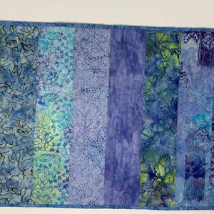 May include: A rectangular, handmade quilt featuring vertical panels of varying blue and green batik fabrics. The panels display floral, leaf, and abstract patterns. The quilt is bordered with a solid blue fabric and measures approximately 24 inches long and 12 inches wide.