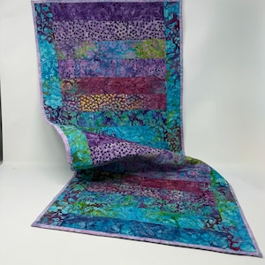 May include: A rectangular fabric table runner featuring a patchwork design of horizontal strips in shades of purple, magenta, teal, and blue. The fabric displays various floral and abstract patterns with a light purple border framing the entire piece.