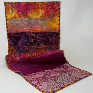 May include: A colorful, quilted table runner with a vibrant mix of pink, orange, yellow, and purple hues. The fabric features floral and abstract patterns, with a contrasting border. The runner is folded, showcasing its layered construction.