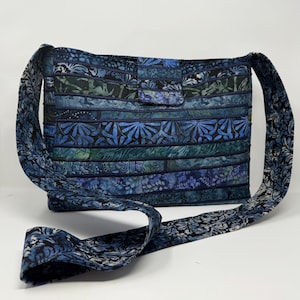 May include: A handmade blue and green quilted shoulder bag. The bag features a rectangular body with a flap closure and a long strap. The fabric has a batik-style pattern with floral and abstract designs. The bag is made of multiple horizontal strips of fabric.