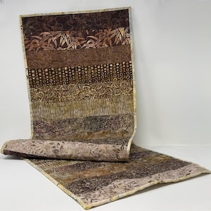 May include: A handmade, rectangular table runner with a patchwork design. The runner features various brown fabrics with leaf and bamboo patterns. The rolled-up section reveals a lighter, floral-patterned fabric. The edges are finished with a cream-colored border.