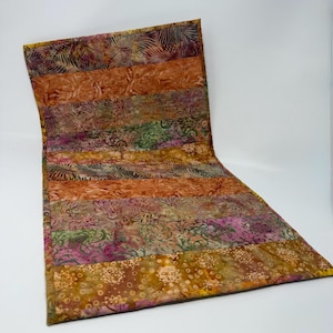 May include: A colorful, quilted table runner with a unique, curved design. The runner features horizontal stripes in shades of orange, brown, green, and pink, with floral and leaf patterns. The edges are finished with a yellow border.