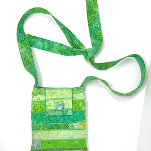 May include: A small green patchwork bag with a long green strap. The bag has a button closure and is made of different shades of green fabric.