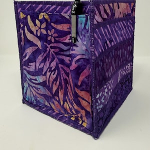 May include: A handmade fabric desk organizer in shades of purple, featuring a floral and leaf pattern. The organizer has multiple pockets and a pen holder with a black pen. The edges are neatly stitched.