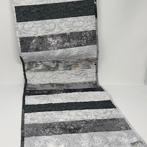 May include: A rectangular quilted table runner featuring alternating horizontal stripes of solid black, mottled gray, and a light gray fabric with a repeating pattern of chickadee birds, pine branches, and pinecones, finished with stippled stitching.