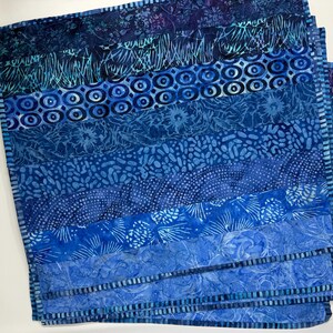May include: A stack of blue fabric squares with various patterns. The designs include circles, floral, and abstract shapes in shades of indigo, teal, and sky blue. The edges are neatly stitched, creating a layered effect.