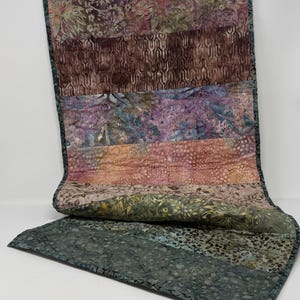 May include: A rectangular fabric table runner featuring horizontal bands of batik-style patterns in shades of purple, mauve, peach, olive green, and dark charcoal grey. The piece is finished with a dark grey binding along all four edges.
