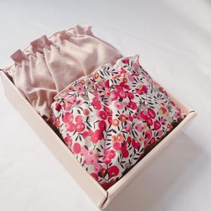 May include: A pink and white floral print fabric is folded and placed inside a pink gift box. The fabric has a ruffled edge.