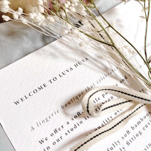 May include: A white card with the text "WELCOME TO LUVA HUVA" is adorned with dried flowers and a cream-colored ribbon with black stripes. The card is from a lingerie boutique.