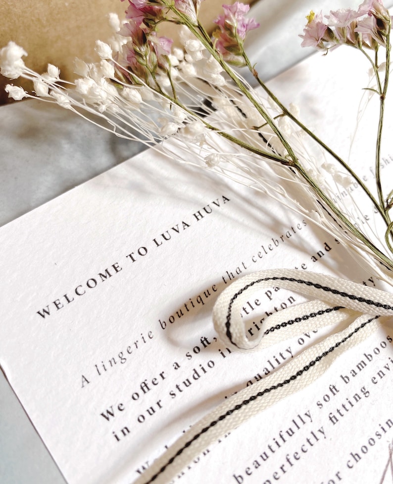 May include: A white card with the text "WELCOME TO LUVA HUVA" is adorned with dried flowers and a cream-colored ribbon with black stripes. The card is from a lingerie boutique.