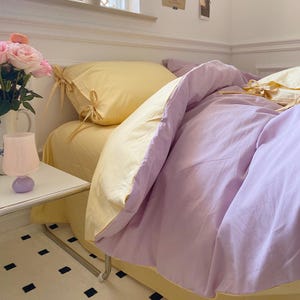 May include: A yellow and purple bedding set with a pillow and a duvet cover. The duvet cover has a yellow border and a purple center. The pillow has a yellow cover with a bow.