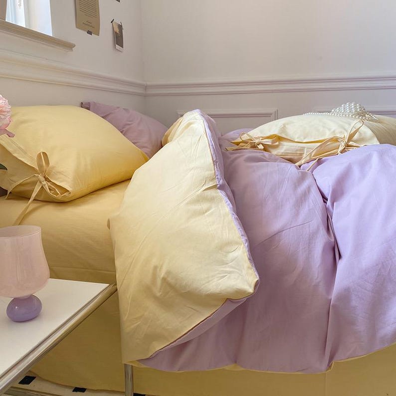 May include: A bed with a yellow and purple duvet cover and matching pillowcases. The duvet cover has a subtle texture and the pillowcases have a simple design. The bed is made with a white sheet and a white pillow.