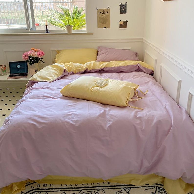 May include: A purple and yellow bed with a yellow pillow and a white decorative accent. The bed is made with a soft, light fabric and has a simple, elegant design.
