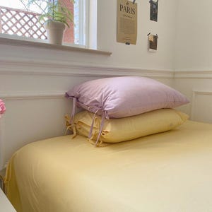 May include: Two pillows on a yellow bedspread. The top pillow is lavender and the bottom pillow is yellow. Both pillows have ties at the corners.