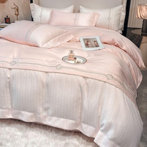 Puede incluir: Un juego de cama rosa y blanco con detalles bordados. El juego incluye una funda nórdica, dos fundas de almohada y una almohada decorativa. La ropa de cama está hecha de tela suave y lujosa y presenta un delicado patrón floral.