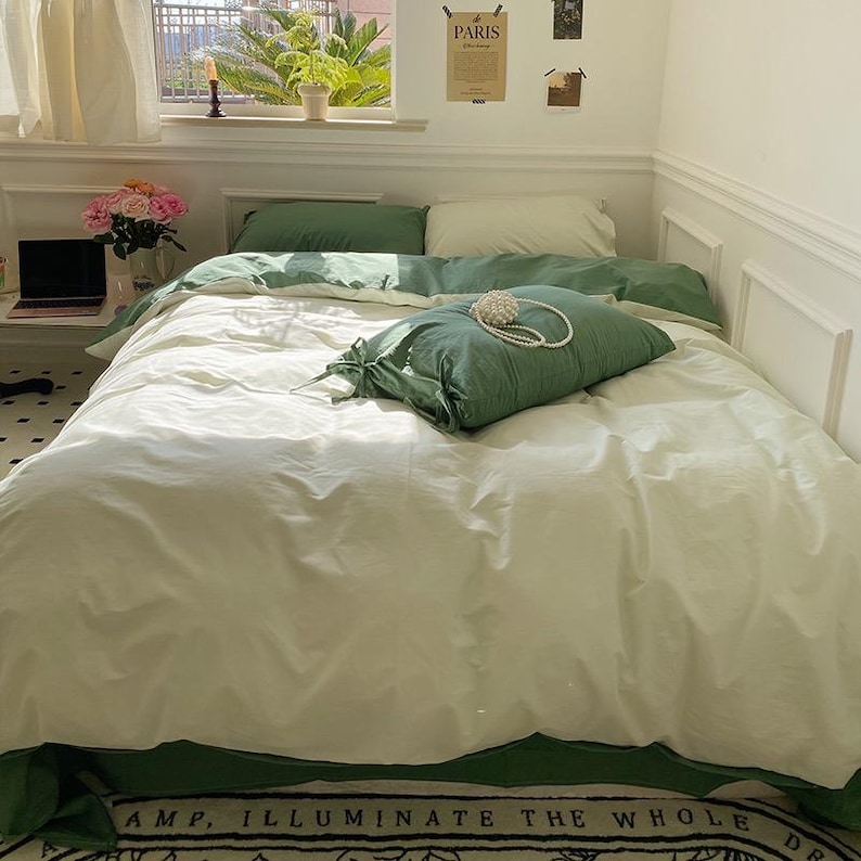 May include: A white bed with green bedding and a green pillow. The pillow has a pearl necklace on it. The bed is in a bedroom with white walls and a window with white curtains.