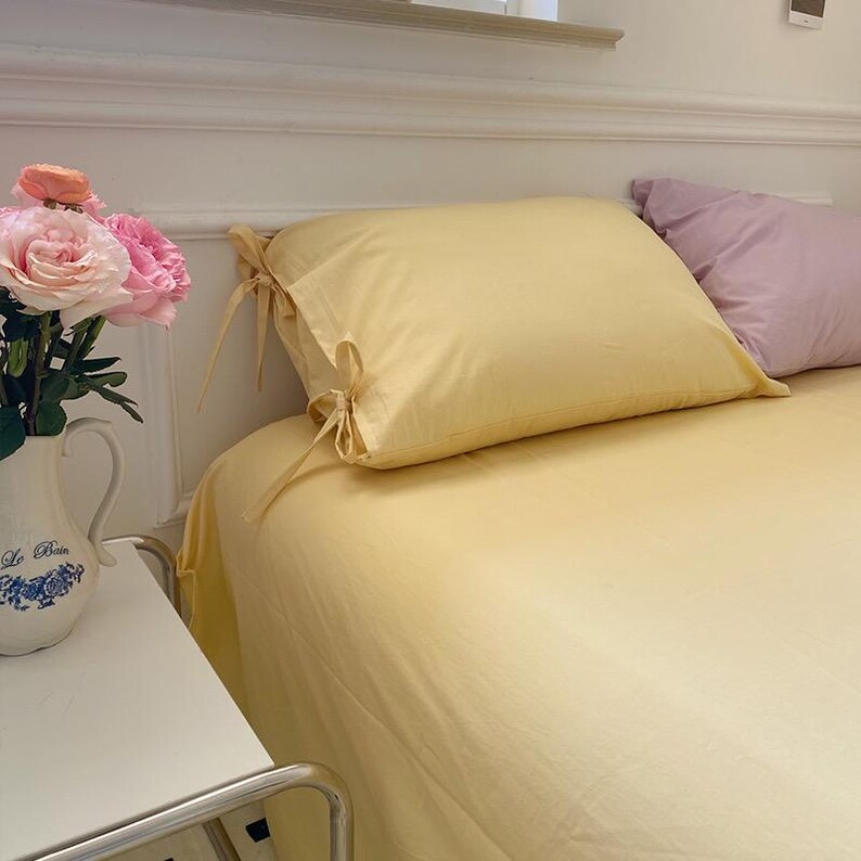 May include: A yellow bed with a yellow pillow with white ties and a purple pillow. The bed is made with crisp, white sheets.