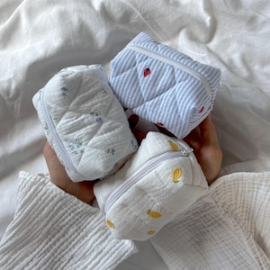 Puede incluir: Tres pequeñas bolsas acolchadas con cremallera hechas de tela suave. Una bolsa presenta rayas azules y blancas con fresas bordadas, una es blanca con patrones florales azules y una es blanca con motivos de limones amarillos bordados.