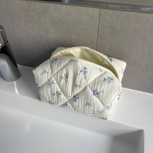 Könnte beinhalten: Eine cremefarbene, gesteppte Kosmetiktasche mit einem Muster aus kleinen blauen Blumen. Die Tasche ist geöffnet und zeigt ein hellgelbes Innenfutter. Sie liegt auf einer weißen Oberfläche, was auf ein Badezimmer hindeutet.