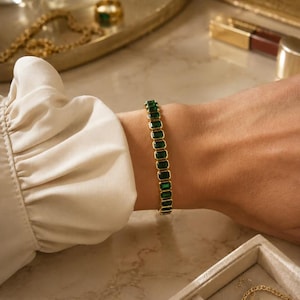 Op de afbeelding: Een gouden armband met rechthoekige smaragdgroene stenen wordt om een pols gedragen. De armband wordt getoond tegen een neutrale achtergrond met andere sieraden en accessoires.