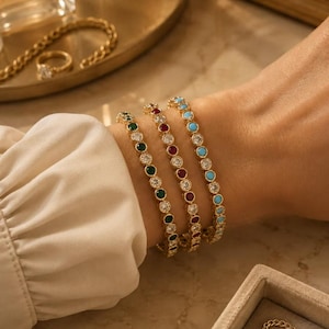 Puede incluir: Tres pulseras doradas con piedras preciosas de colores se muestran en una muñeca. Las pulseras presentan piedras verdes, rojas y turquesas, intercaladas con cristales transparentes. El fondo es neutro.