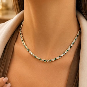 Pode incluir: Colar dourado com detalhes de gemas redondas de esmeralda e transparentes alternadas. O colar é usado ao redor do pescoço, com uma vista de perto que destaca os detalhes da joia. As gemas são engastadas em um design delicado e elegante.