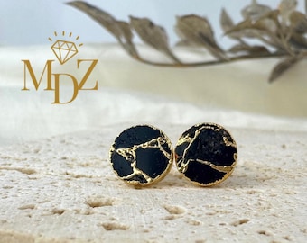 May include: A pair of round stud earrings. Each earring features a black surface with gold-colored veining and a gold-colored border. The earrings are displayed on a textured, light-colored surface. The logo "MDZ" is visible in the upper left corner.