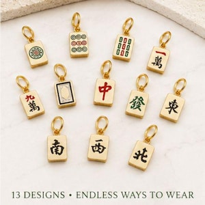 Op de afbeelding: Dertien goudkleurige metalen bedels in de vorm van rechthoekige Mahjong-tegels, elk voorzien van een uniek gekleurd emaille symbool of Chinees karakter en een kleine ronde ring aan de bovenkant. De tekst 13 DESIGNS &bull; ENDLESS WAYS TO WEAR is zichtbaar.