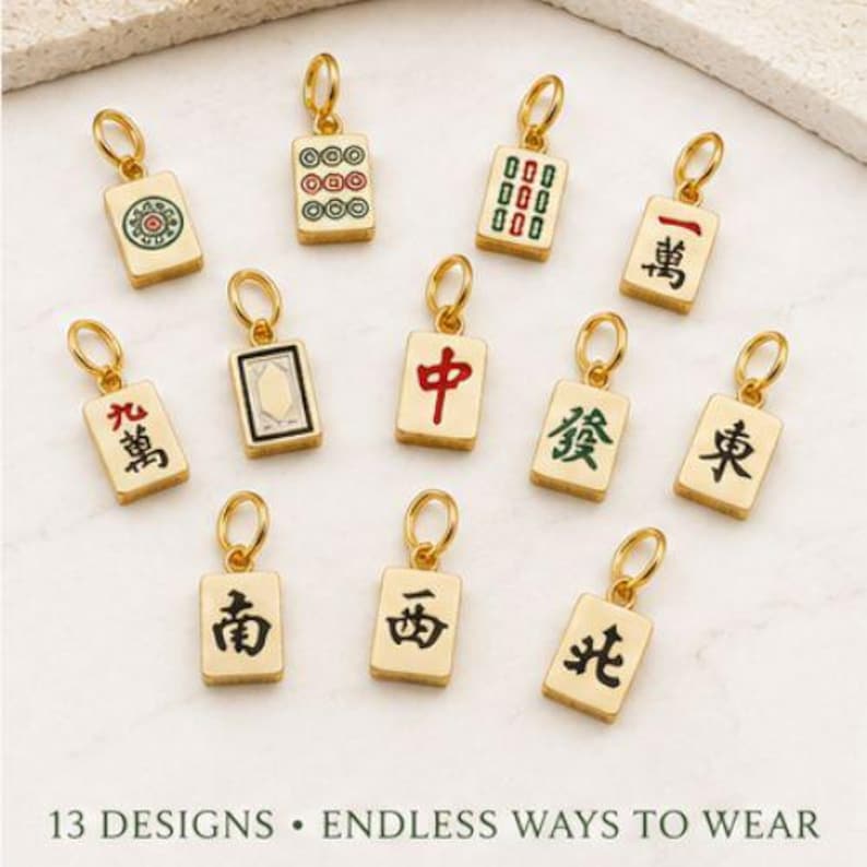 Op de afbeelding: Dertien goudkleurige metalen bedels in de vorm van rechthoekige Mahjong-tegels, elk voorzien van een uniek gekleurd emaille symbool of Chinees karakter en een kleine ronde ring aan de bovenkant. De tekst 13 DESIGNS &bull; ENDLESS WAYS TO WEAR is zichtbaar.