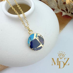 May include: A gold-colored chain necklace with a circular pendant. The pendant features a mosaic design with shades of blue, navy, and gold. The necklace is displayed against a white background, with a logo in the bottom right corner.