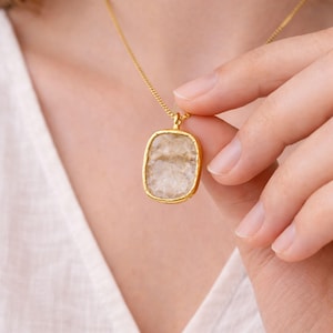 May include: A gold-colored necklace with a rectangular pendant. The pendant has a light-colored, textured stone set within a gold frame. The necklace is being held up by a person's hand, showcasing the jewelry's design and color.