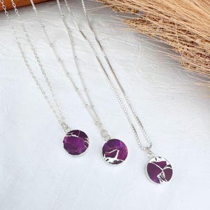 May include: Three silver necklaces with round, purple pendants. The pendants have a cracked, mosaic-like design. The necklaces are displayed on a white surface, with dried plants in the background.