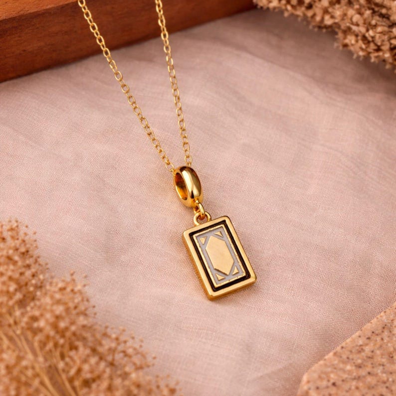 Op de afbeelding: Een goudkleurige ketting met een rechthoekige hanger met een centraal geometrisch ontwerp. De hanger heeft een gepolijst gouden frame rondom een verdiept gedeelte met een lichtgekleurde inleg en een donkere rand, hangend aan een fijne ketting.