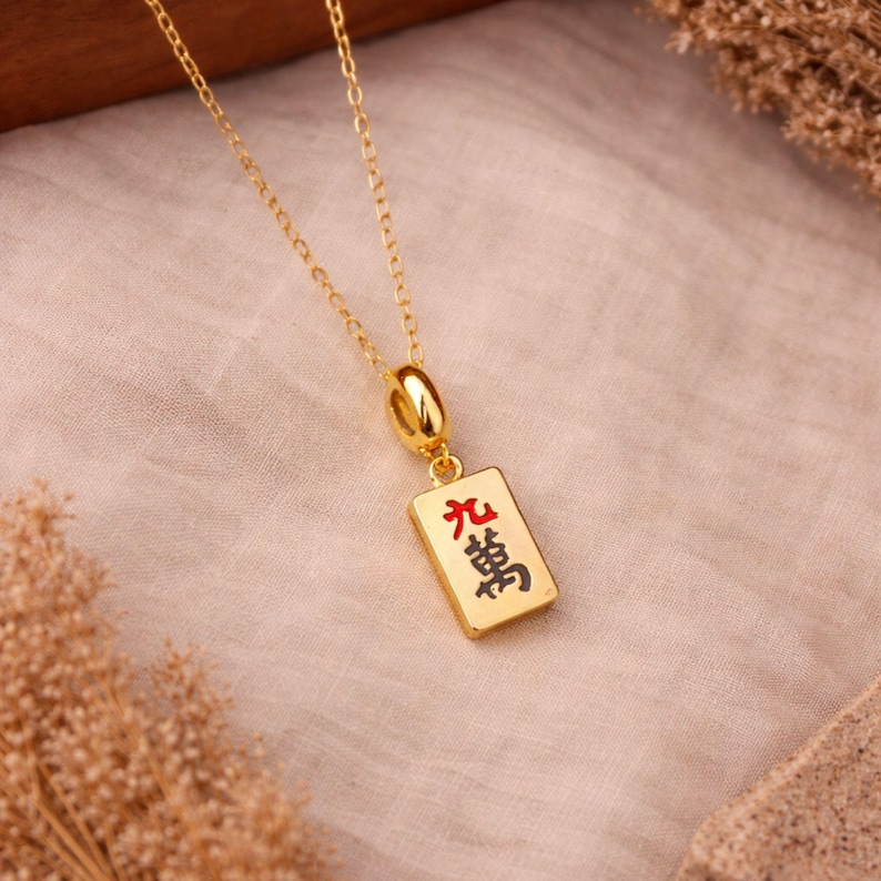 Op de afbeelding: Een goudkleurige ketting met een rechthoekige hanger in de vorm van een mahjongtegel. De hanger toont het Chinese karakter voor negentienduizend in zwarte inkt met een rood accent, hangend aan een fijne goudkleurige ketting.