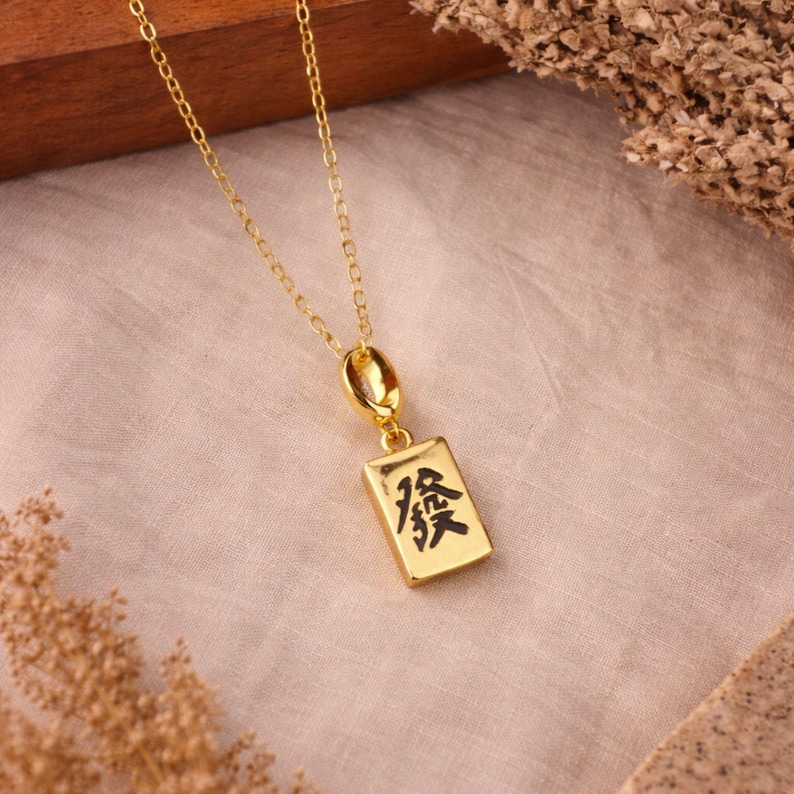 Op de afbeelding: Een goudkleurige ketting met een rechthoekige hanger in de vorm van een mahjongtegel. De hanger toont het zwarte Chinese karakter voor 'Fa' of 'geluk' op de voorzijde en hangt aan een fijne goudkleurige schakelketting.