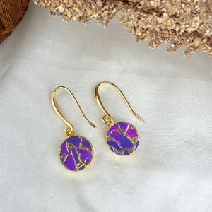 May include: A pair of gold-colored drop earrings. Each earring features a round, purple and gold-colored pendant. The purple pendant has a mosaic design. The earrings are displayed on a white surface.