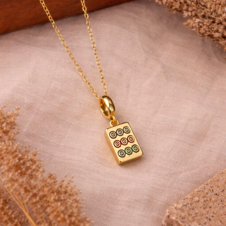 Op de afbeelding: Een goudkleurige ketting met een rechthoekige hanger in de vorm van een mahjongsteen. De hanger toont drie rijen cirkelvormige symbolen in zwart, rood en groen emaille, hangend aan een fijne goudkleurige ketting met een gepolijste afwerking.