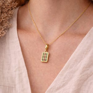 Op de afbeelding: Een gouden ketting met een rechthoekige hanger. De hanger heeft een gouden rand en een centrale inleg met drie verticale rijen van kleine, afwisselend rode en groene rechthoekige vormen op een witte achtergrond.