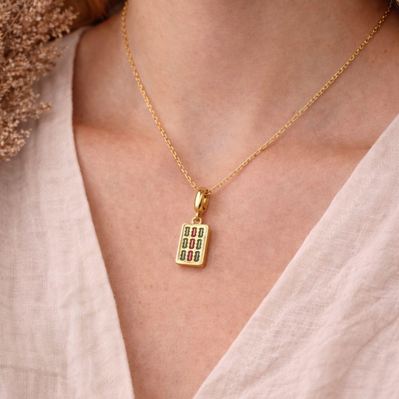 Op de afbeelding: Een gouden ketting met een rechthoekige hanger. De hanger heeft een gouden rand en een centrale inleg met drie verticale rijen van kleine, afwisselend rode en groene rechthoekige vormen op een witte achtergrond.