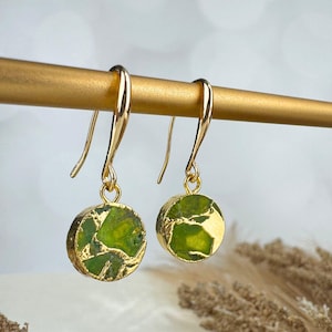 May include: A pair of gold-colored drop earrings. Each earring features a round, green stone with gold-colored accents, set in a gold-colored bezel. The earrings hang from gold-colored ear wires.