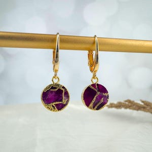 May include: A pair of gold-colored hoop earrings with round, deep purple pendants. The pendants have a gold-colored, cracked-stone design. The earrings are displayed on a gold-colored bar against a blurred background.
