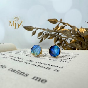 May include: Two round stud earrings with a blue and green iridescent stone set in gold-colored metal. The earrings are displayed on an open book with text. The MDZ logo is visible in the upper left corner.