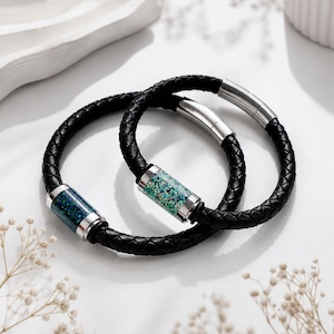 May include: Two braided black leather bracelets featuring cylindrical metal accents. One bracelet contains a dark blue and green speckled inlay, while the other features a light blue and white speckled inlay. Both have silver-toned metal clasps.